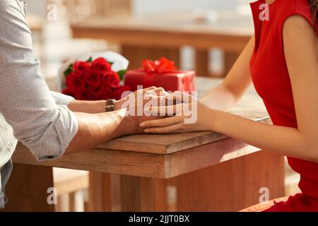 Romantic date: unrecognizable young woman holding hands with her boyfriend while sitting at wooden table, gift box and bouquet of red roses seen behind them Stock Photo