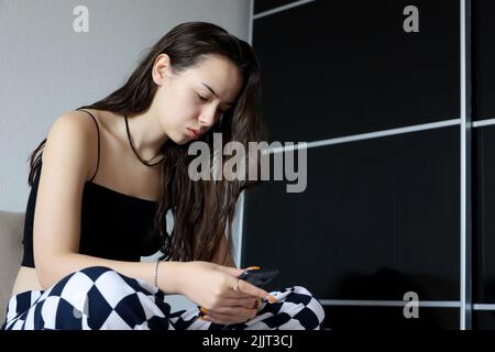 Pretty girl with long hair sitting with smartphone on sofa. Online communication, leisure at home Stock Photo