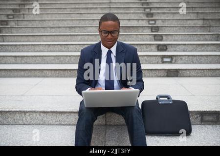 Cheerful African-American business executive sitting outdoors and working on laptop Stock Photo