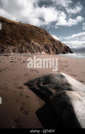 Vertical shot from the sandy shore to a steep cliff by the sea and footprints in the sand under a blue cloudy sky Stock Photo