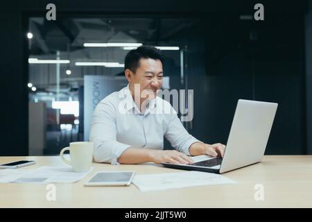 Young handsome male Asian freelancer conducts a video meeting on a video call from a laptop. Sitting at the table in a modern office, smiling, talking. Stock Photo