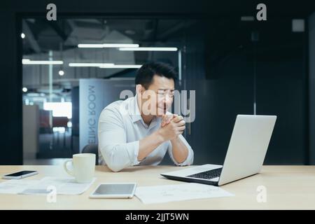 Young handsome male Asian freelancer conducts a video meeting on a video call from a laptop. Sitting at the table in a modern office, smiling, talking. Stock Photo