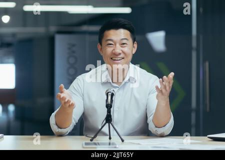 Portrait of a young handsome Asian man speaking to the camera into a microphone, recording a video message, conducting a webinar, making a podcast, gesturing with his hands, smiling in office Stock Photo