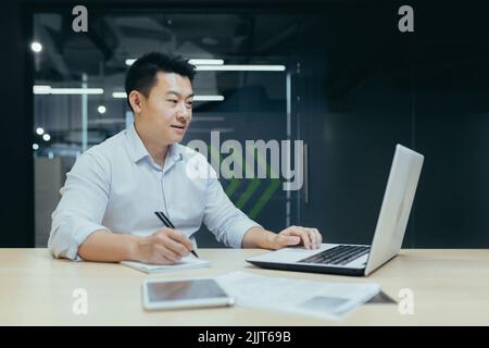 Young businessman, freelancer, Asian working with documents, writing with a pen, working on a laptop Sitting at a desk in a modern office Stock Photo