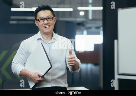 Portrait of a young Asian man intern, assistant in the office. He is standing in glasses, pointing ok, holding a folder in his hands, smiling, looking at the camera. Stock Photo