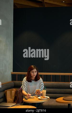 Asian young woman writing plans for the day and eating breakfast in cafe Stock Photo