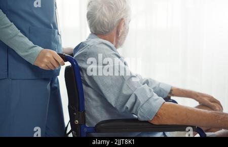 Nurse putting wheelchair with senior man  window. Unhappy senior man having geriatric or depression disease. Therapist taking care, giving support and Stock Photo