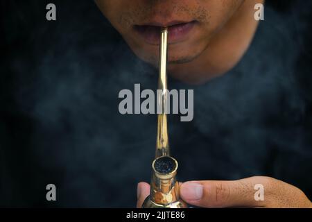 Asian man smokes marijuana from a pipe at home. Studio shoot with model simulating smoking pot with a pipe in a dark background. Cannabis legalisation Stock Photo