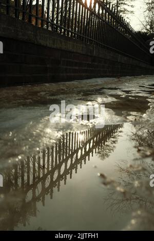 A vertical of a puddle on a pavement covered with snow reflecting metallic fence in Minnesota in sunlight Stock Photo