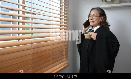 Smiling senior female judge in robe gown uniform having phone conversation while standing in her personal office Stock Photo