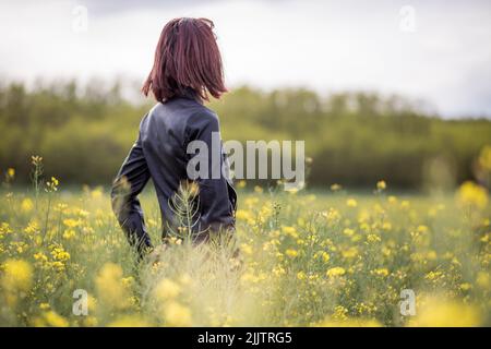 A back shot portrait of a young woman standing among blooming yellow flowers in the field on a sunny day Stock Photo