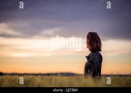 A beautiful portrait of a young woman standing among long green grass in the field against dusk sky at sunset Stock Photo