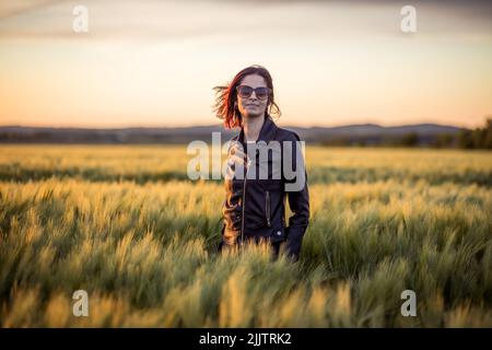 A beautiful portrait of a young Caucasian woman standing among long green grass in the field against dusk sky at sunset Stock Photo