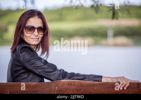 A beautiful portrait of a young Caucasian woman sitting on a wooden bench by the lake in the park and looking at the camera on a sunny day Stock Photo