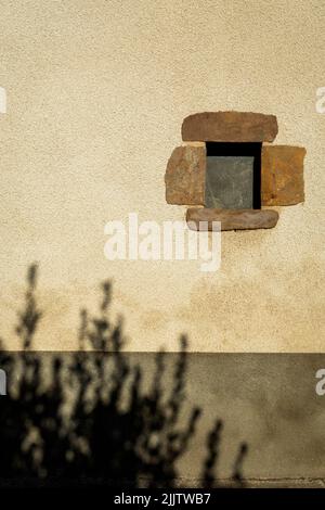 A vertical shot of a small square window on a beige wall with the silhouette of a bush Stock Photo