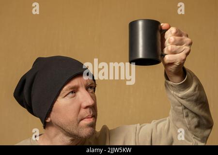 A man in a black hat looks at a black cup in his hand. Portrait of a middle-aged European man, close-up Stock Photo