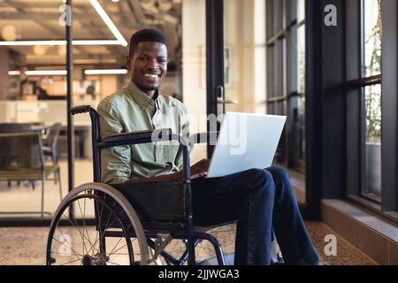 Smiling african american businessman with disability using laptop in wheelchair at workplace Stock Photo