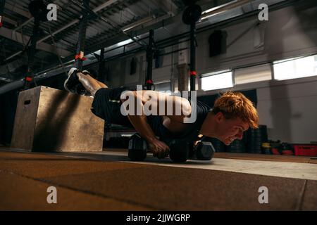 Portrait of young sportive red-headed man doing push up exercises with dumbbells isolated on indoor gym background Stock Photo