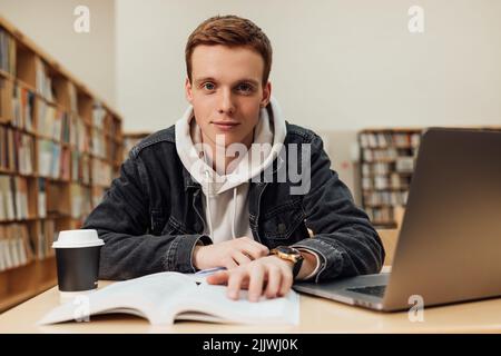 Portrait of a young man sitting at desk in library and looking at camera Stock Photo