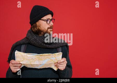 Ginger puzzlement man wearing hat and scarf examining map isolated over red background Stock Photo