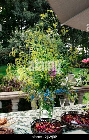 a bouquet of wild flowers with herbs in a vase on a table on a summer terrace in the garden. Festive table with vegetarian dishes at the party. Outdoo Stock Photo
