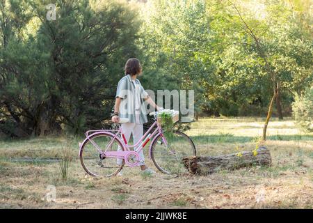 Side view of unrecognizable back looking young long haired man holding a pink vintage cute bike in the park Stock Photo