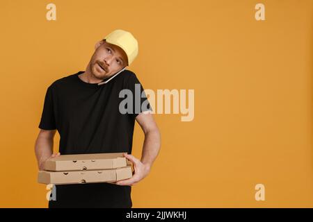 White delivery man with pizza boxes talking on mobile phone isolated over yellow background Stock Photo
