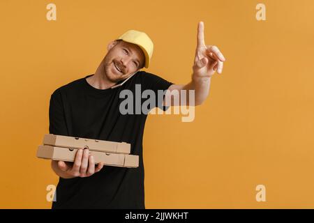 White delivery man with pizza boxes talking on mobile phone isolated over yellow background Stock Photo
