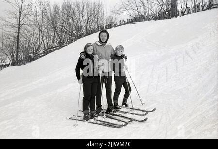 1966, historical, wintertime, snow on the ground and outside on a hillside, a mother with her two young children standing together for a photo in their skis and outdoor clothing, England, UK. Stock Photo