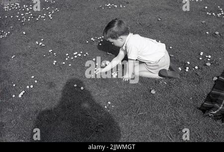 1960s, historical, springtime and outside on a field, a young boy, kneeling down on hands and knees on a grass field, picking daisy petals from the flowers, England, UK. An April birth flower, daisies are said to symbolise innocence and hope. In Britain, the flower is known as the common daisy or lawn daisy. Stock Photo