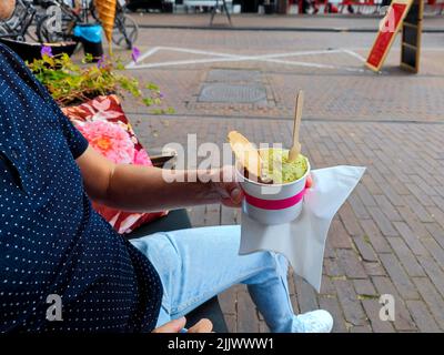 White male hand holds a cup with scoops of ice cream on a bench next to an ice cream shop. There are no recognizable persons or trademarks in the shot Stock Photo