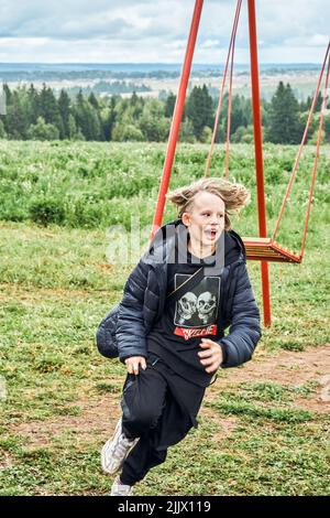 Cheerful boy runs after jumping off swings against lush trees and green grass in countryside. Cute schoolboy with long blond hair on nature. No logo Stock Photo