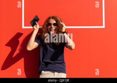 Smiling young Woman with headphones and smartphone against orange wall Stock Photo