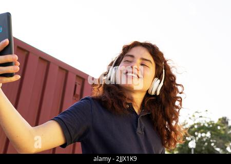 Smiling young Woman with headphones taking selfie through smartphone against sky Stock Photo