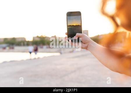 Cropped hand of young Woman photographing beach through smartphone during sunset Stock Photo