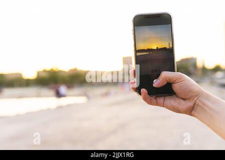 Cropped hand of young Woman photographing beach through smartphone during sunset Stock Photo