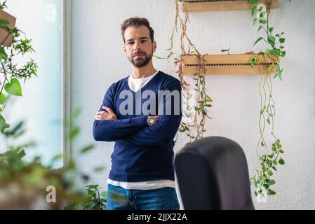 Serious bearded male employee in stylish wear looking at camera while standing in light modern workspace with blurred green leaves Stock Photo