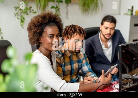 Positive African American female worker looking at camera while sitting near group of colleagues against blurred background in light office Stock Photo