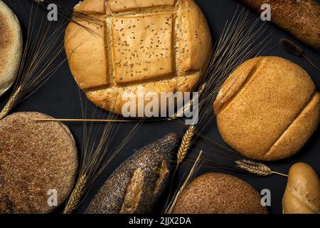 Top view background of assorted bread loaves and baguette placed near heap of wheat spikes on black surface in studio Stock Photo