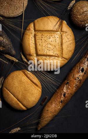 Top view background of assorted bread loaves and baguette placed near heap of wheat spikes on black surface in studio Stock Photo
