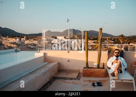 Front view of young bearded male traveler in white robe and sunglasses relaxing on lounger on rooftop and messaging on smartphone against sunset sky d Stock Photo