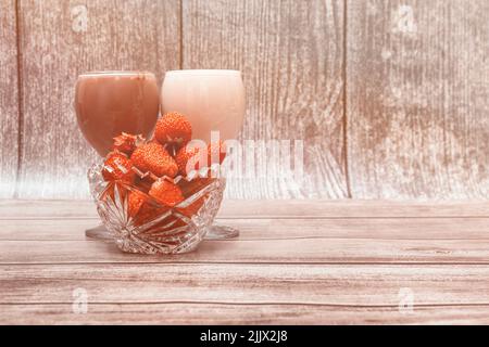 Vase with ripe strawberries and two glasses with multi-colored fruit cocktails stand on wooden table surface and against wooden wall. Stock Photo