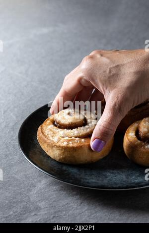 Cropped unrecognizable person holding cinnamon roll bun, Kanelbulle traditional Swedish dessert served in black plate Stock Photo