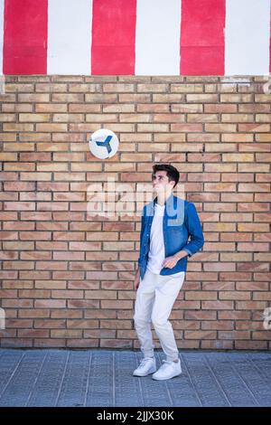Full length of young man looking at football in mid-air while standing against brick wall Stock Photo