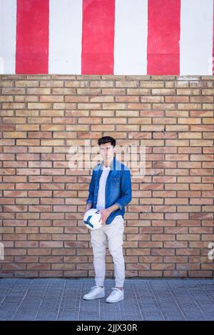 Full length portrait of handsome young man standing with football while standing against brick wall Stock Photo