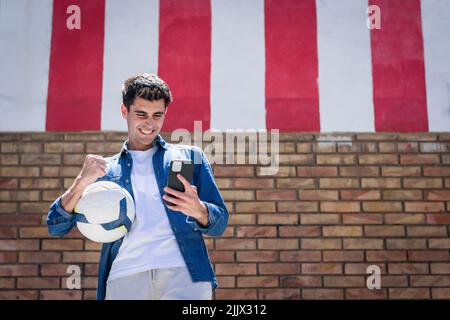 Cheerful young man with football using smartphone while clenching fist Stock Photo