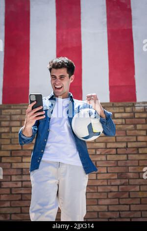 Cheerful young man with football using smartphone while clenching fist Stock Photo