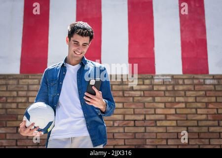 Cheerful young man with football using smartphone standing near brick wall Stock Photo