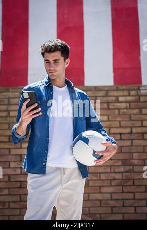 Young man with football using smartphone standing near brick wall Stock Photo
