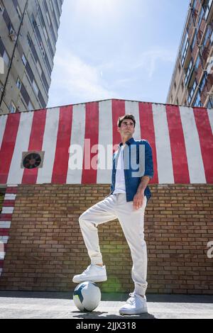 From below young man with foot over football standing on footpath looking away Stock Photo